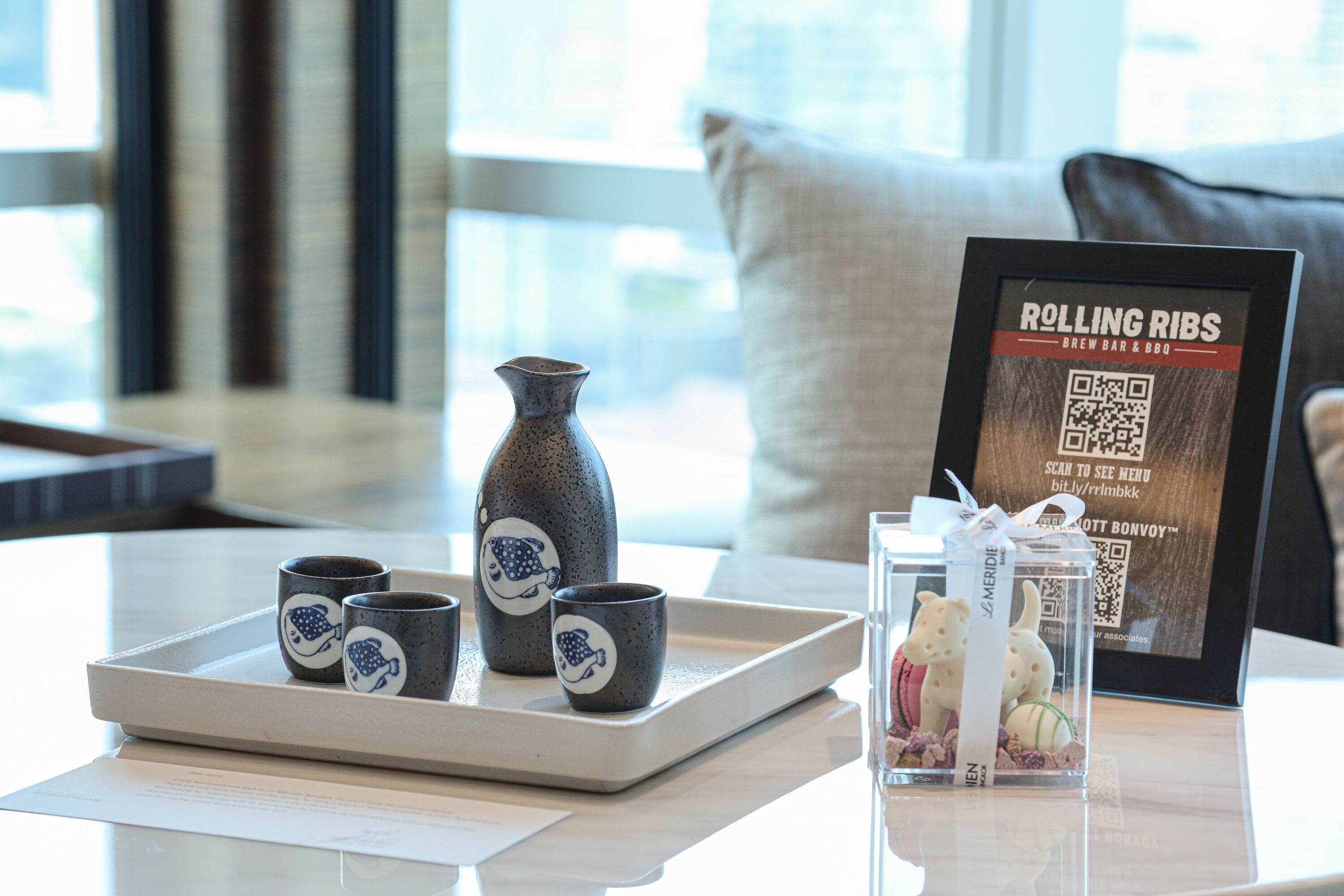 Un plateau avec trois petites tasses et un vase posé sur une table, un menu encadré et un verre décoré d’un ours en peluche.