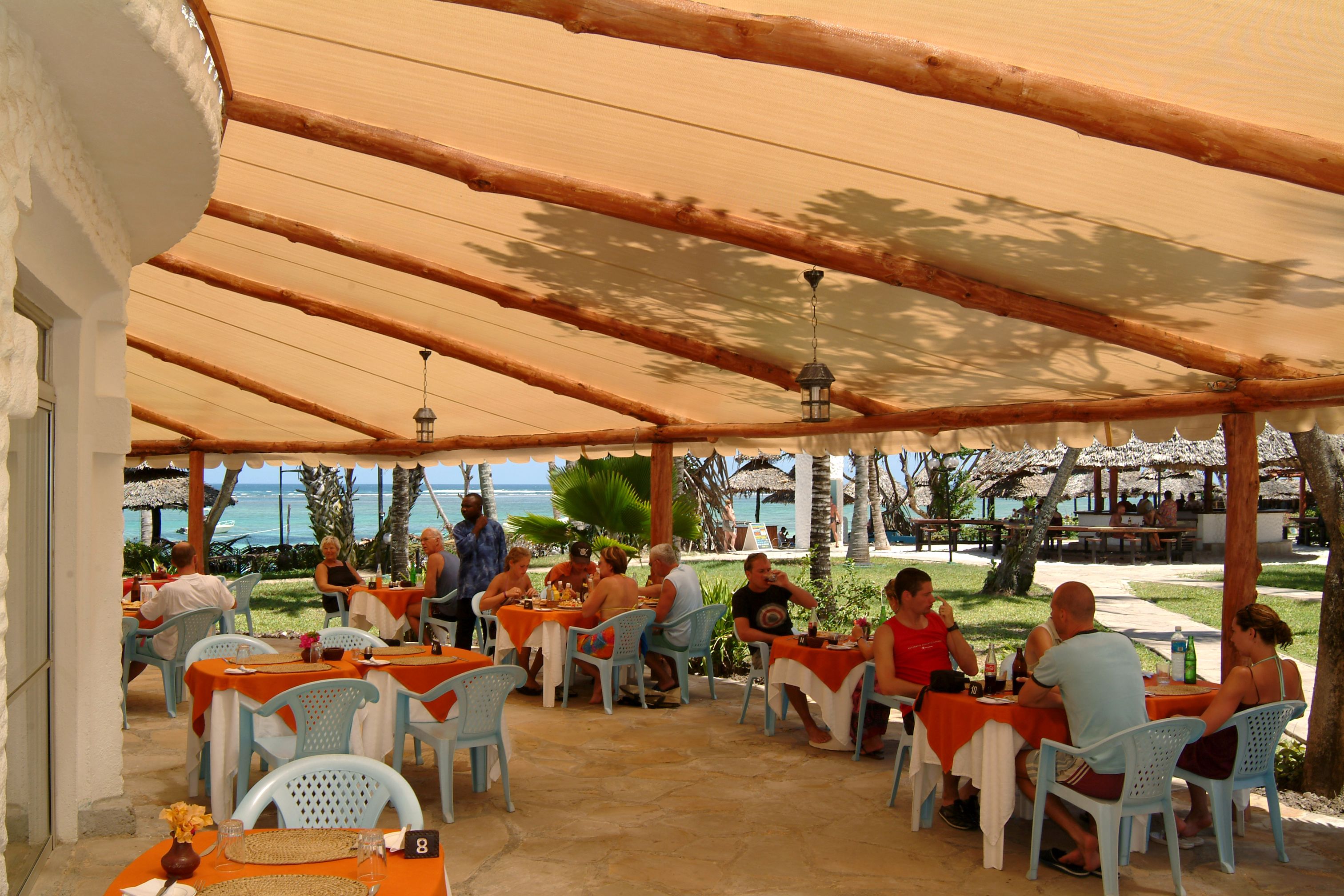 Un área de comedor al aire libre con mesas y sillas bajo un toldo de madera, con vista al océano.