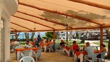 An outdoor dining area with tables and chairs under a wooden canopy, overlooking the ocean.