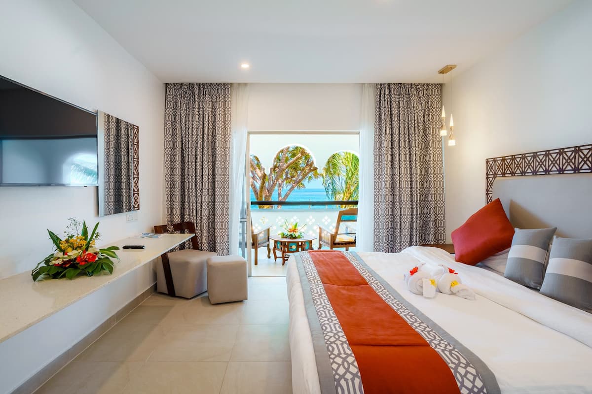 Ein Hotelzimmer mit einem Bett, einem Schreibtisch, einem Stuhl, einem kleinen Tisch mit Blumen und Blick auf das Meer durch ein großes Fenster.