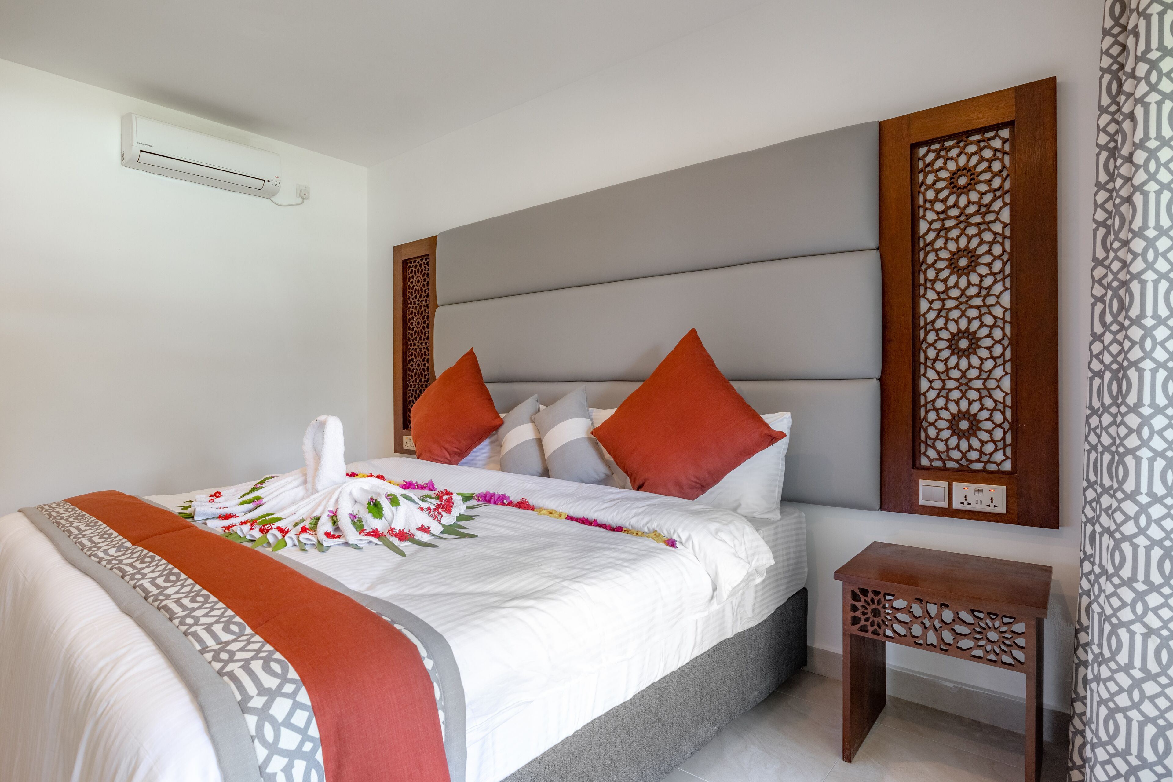 Una cama bien hecha con edredón blanco, almohadas naranjas y una toalla decorativa. Hay una mesita de noche de madera al lado de la cama.