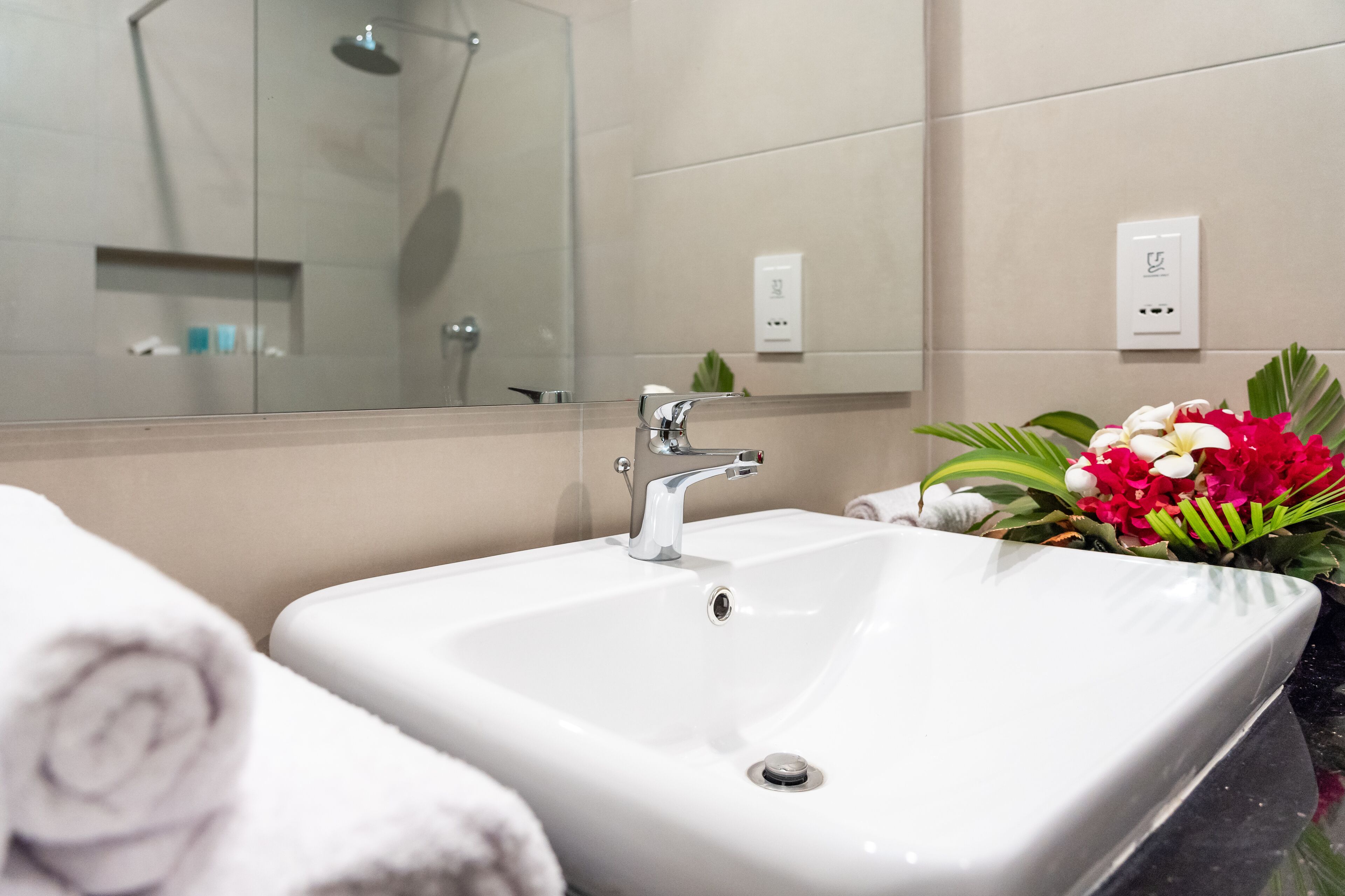 Un baño con lavabo blanco, un espejo y ducha. Hay toallas y un ramo de flores en la encimera.