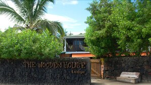 Un muro de piedra con el cartel de 'The Wooden House Lodge', un portón de madera y una palmera.