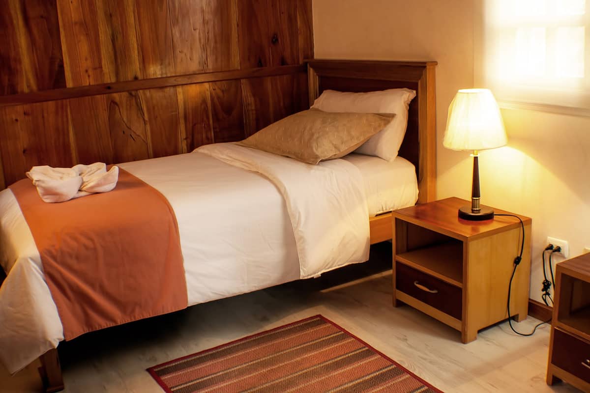 Una cama bien hecha con un cabecero de madera, una lámpara de noche y una mesita de noche de madera con un cajón.