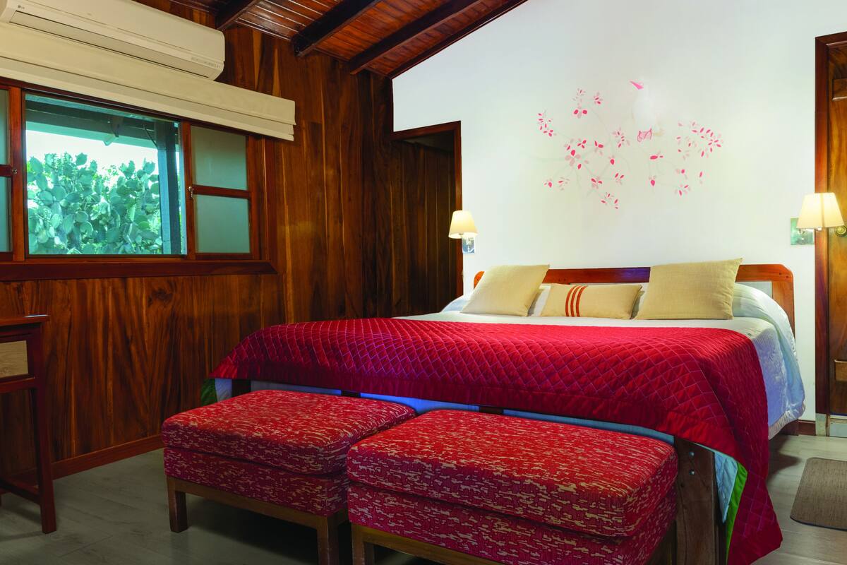 Una habitación de madera con una cama, dos lámparas de noche, un ventanal con vistas y una pared decorada con motivos florales.
