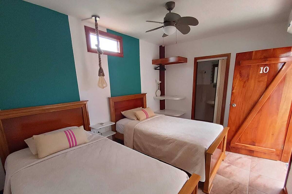 Habitación de hotel con dos camas, ventilador de techo, ventana, puerta de madera con el número 10 y zona de ducha.