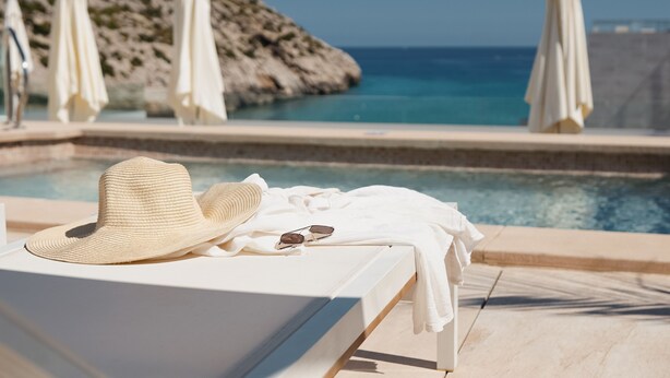 Una tumbona con un sombrero y gafas de sol, vista a la piscina y sombrillas.