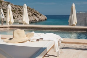 Una tumbona con un sombrero y gafas de sol, vista a la piscina y sombrillas.