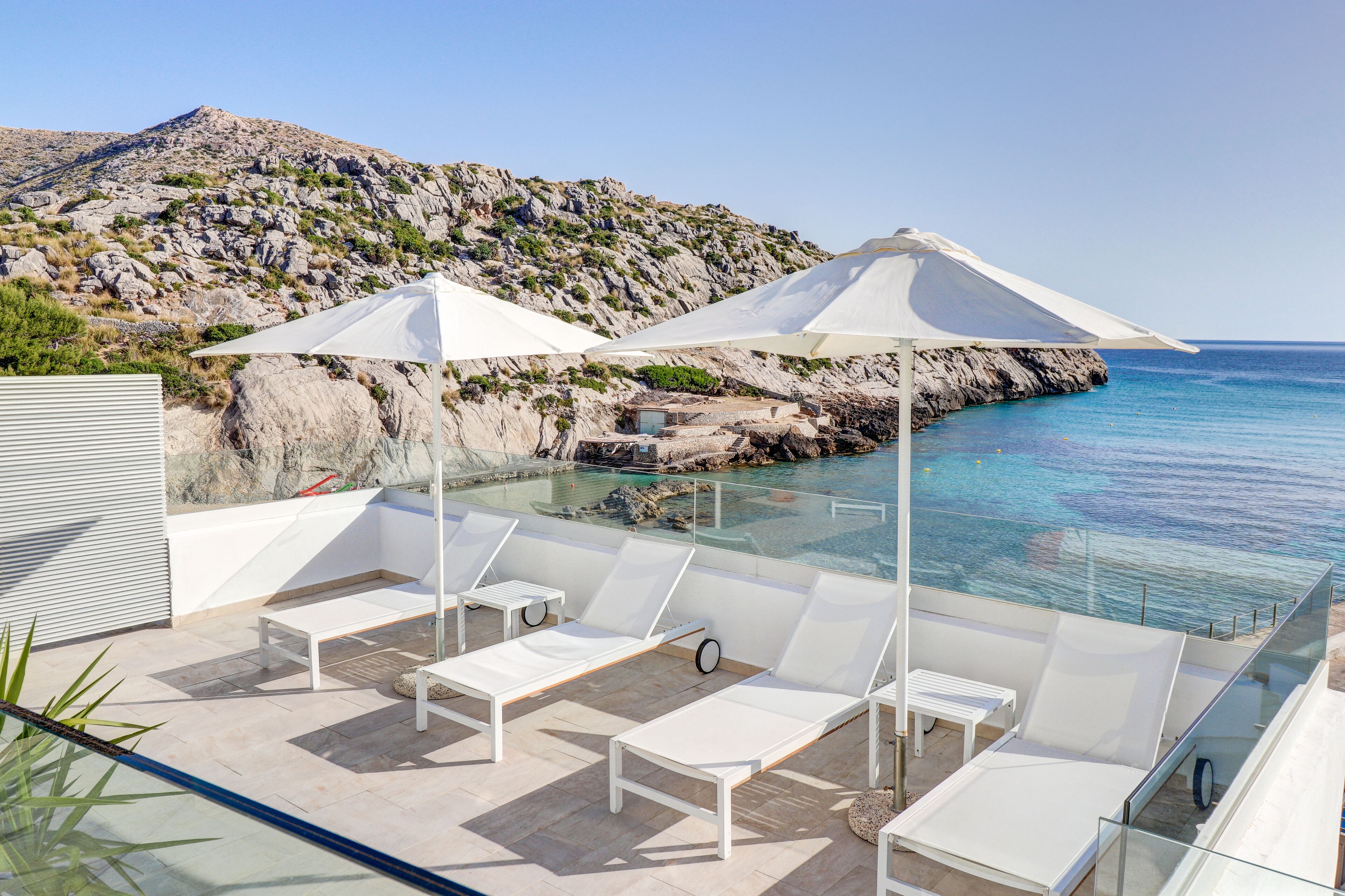 A rooftop terrace with white lounge chairs and umbrellas, overlooking a rocky coastline and clear blue sea.