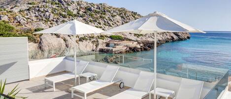 A rooftop terrace with white lounge chairs and umbrellas, overlooking a rocky coastline and clear blue sea.
