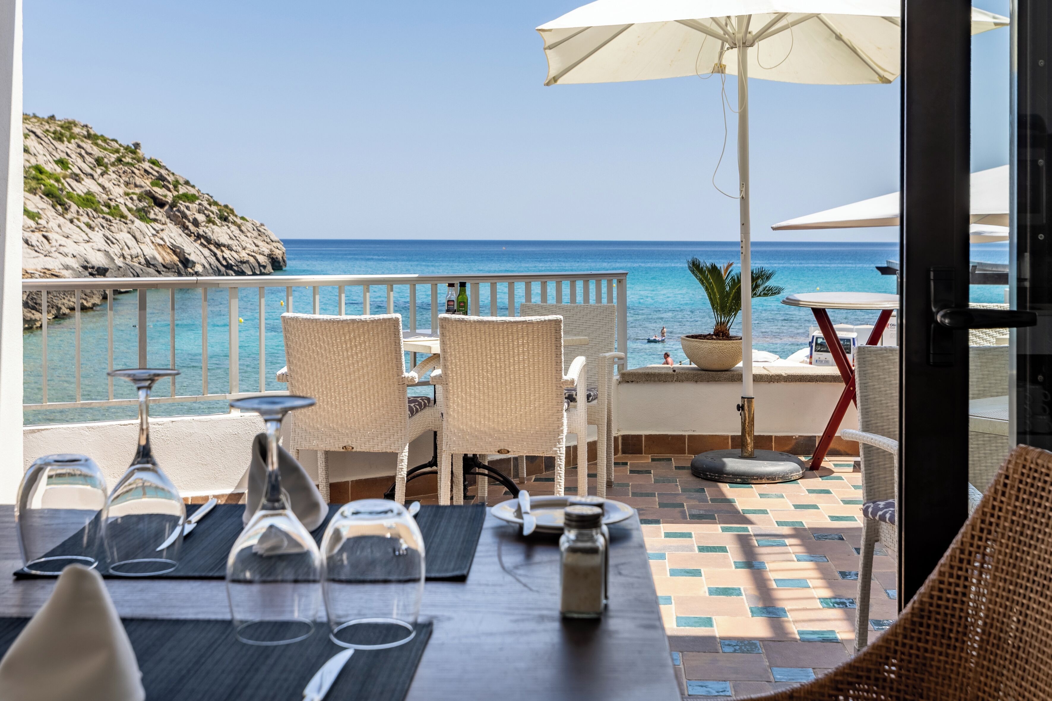 A terrace with wicker chairs, a table set for dining, a potted plant, and a view of the sea.