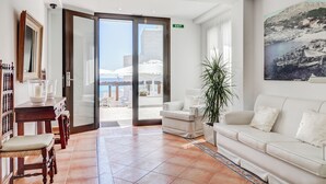 Un vestíbulo de hotel con un sofá blanco, una mesa de madera con sillas, una planta grande en maceta y vistas al mar a través de un ventanal.