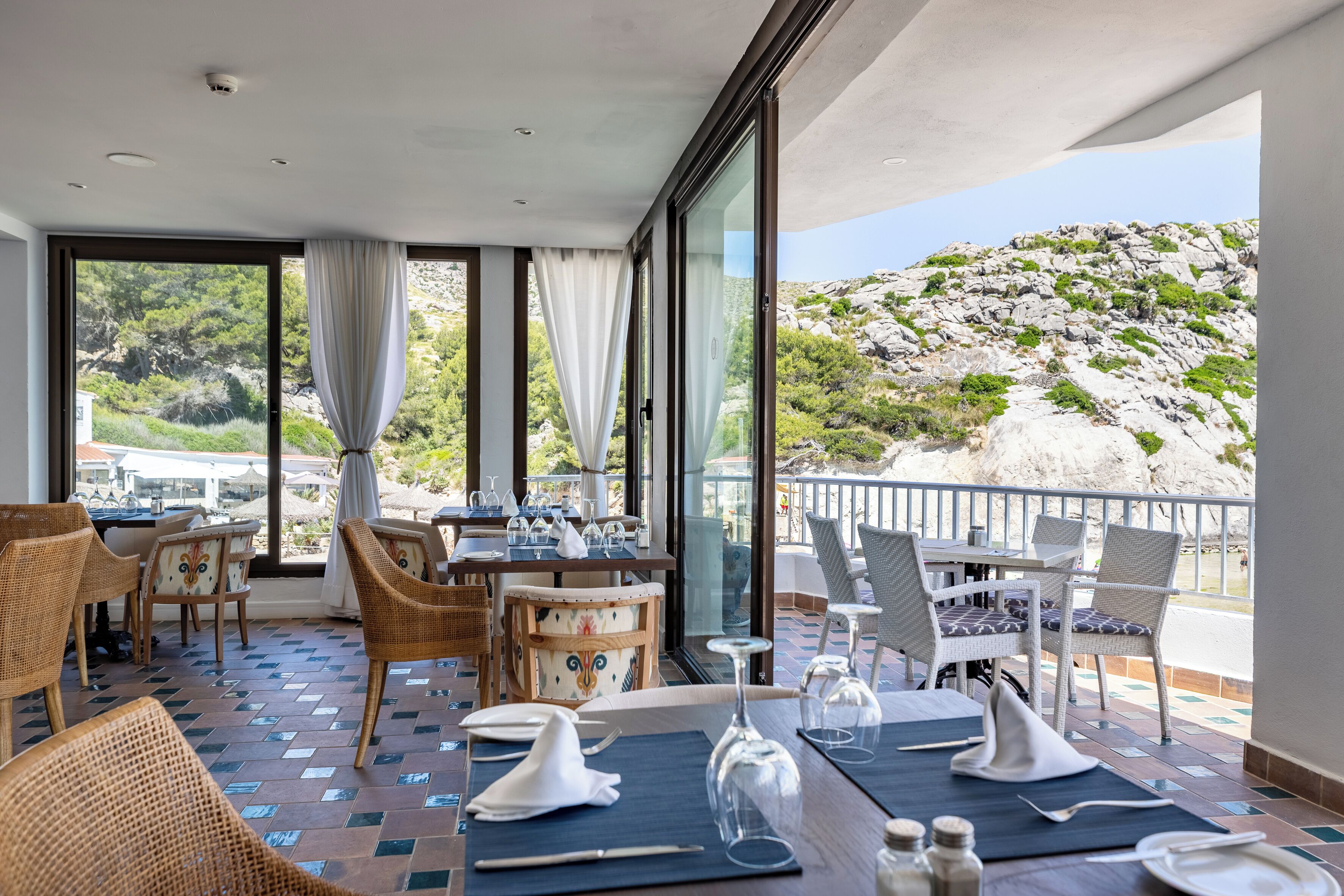 A restaurant with outdoor seating, tables set with white napkins and wine glasses, and a view of a rocky hillside.
