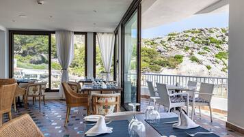 A restaurant with outdoor seating, tables set with white napkins and wine glasses, and a view of a rocky hillside.