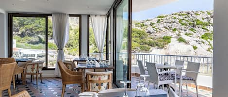 A restaurant with outdoor seating, tables set with white napkins and wine glasses, and a view of a rocky hillside.