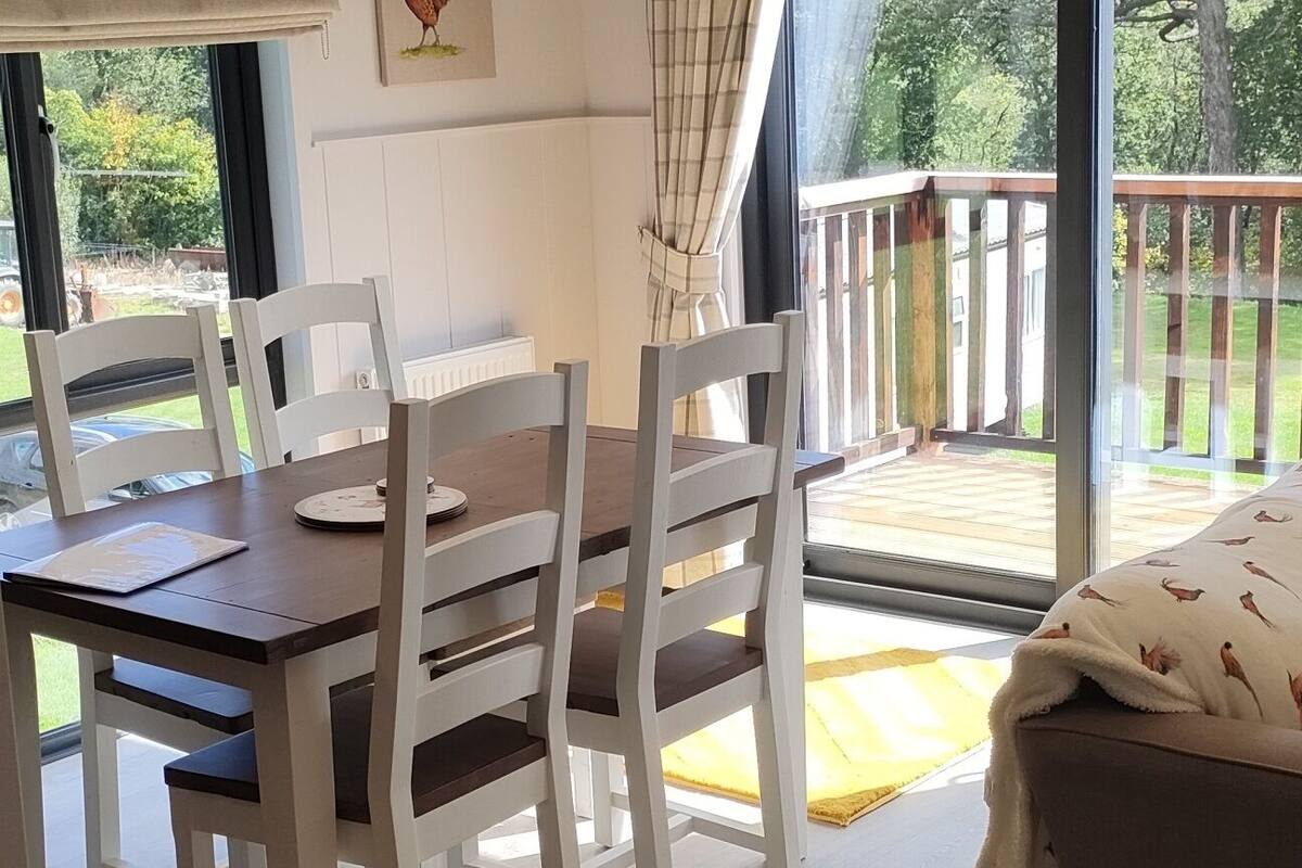 A dining area with a wooden table and white chairs, a large window with a view of a deck, and a ceiling with recessed lighting.