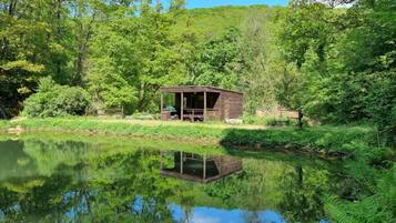 A wooden cabin with a deck, surrounded by lush greenery and a calm pond.