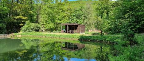 A wooden cabin with a deck, surrounded by lush greenery and a calm pond.