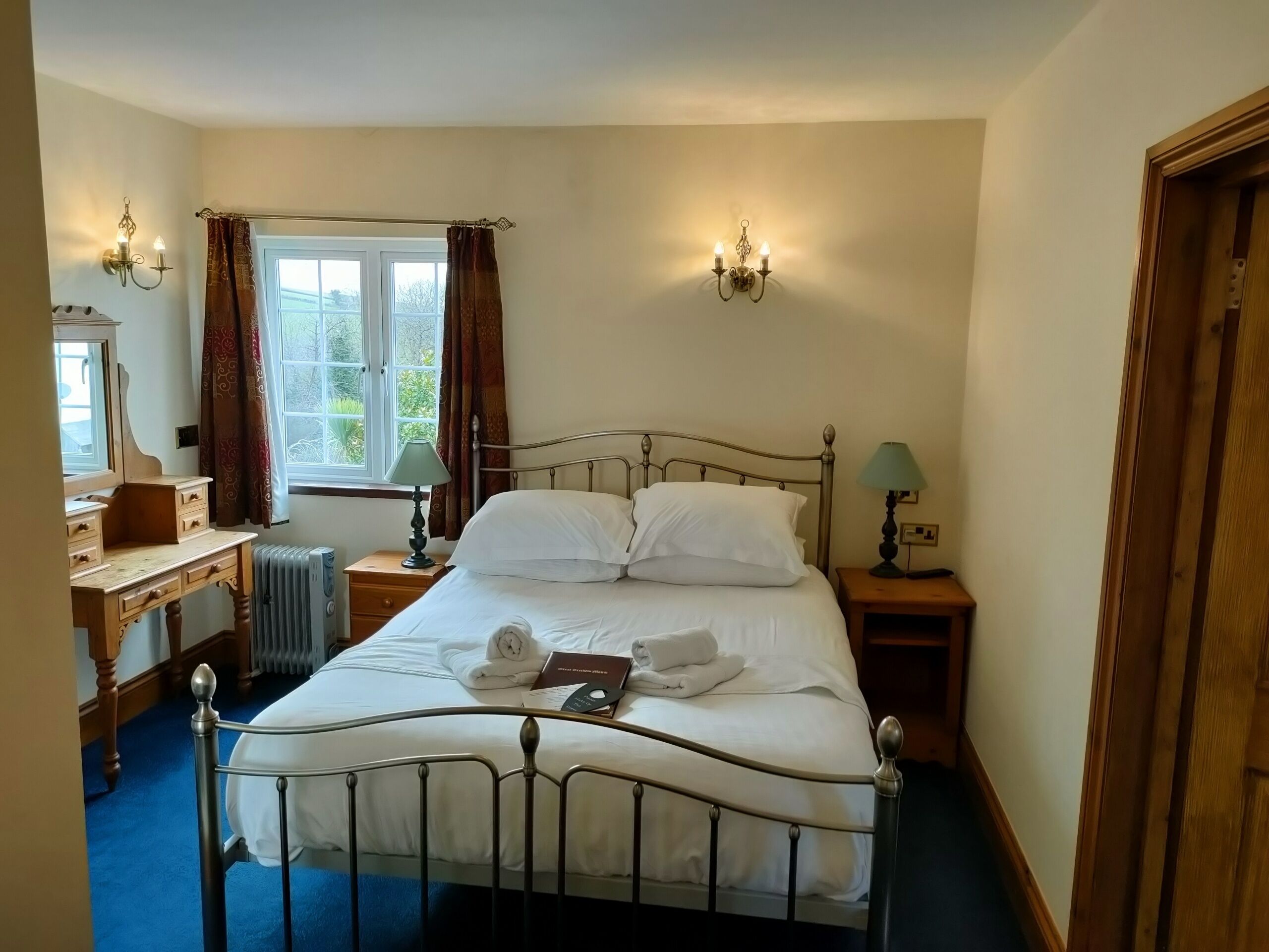 A neatly made bed with white linens and towels, a wooden bedside table with a lamp, a dresser with a mirror, and a window with curtains.