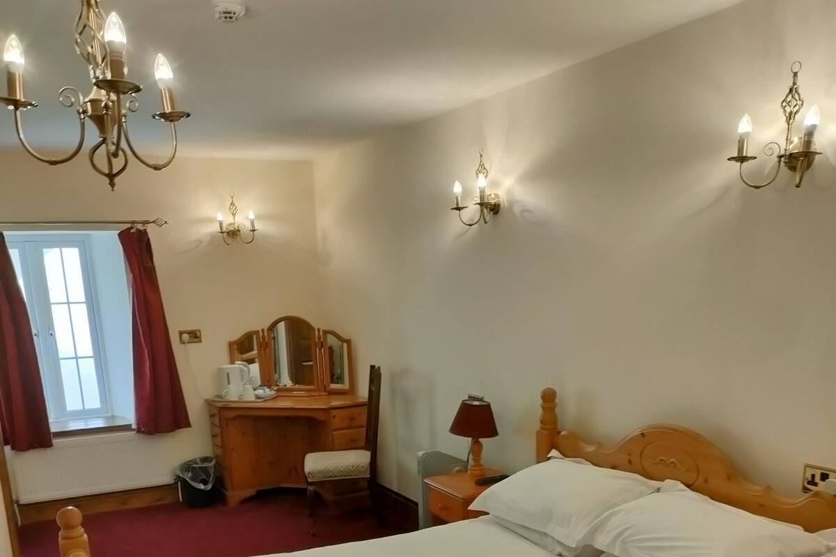 A hotel room with a wooden bed, a wooden desk, a chair, a lamp, and a chandelier.