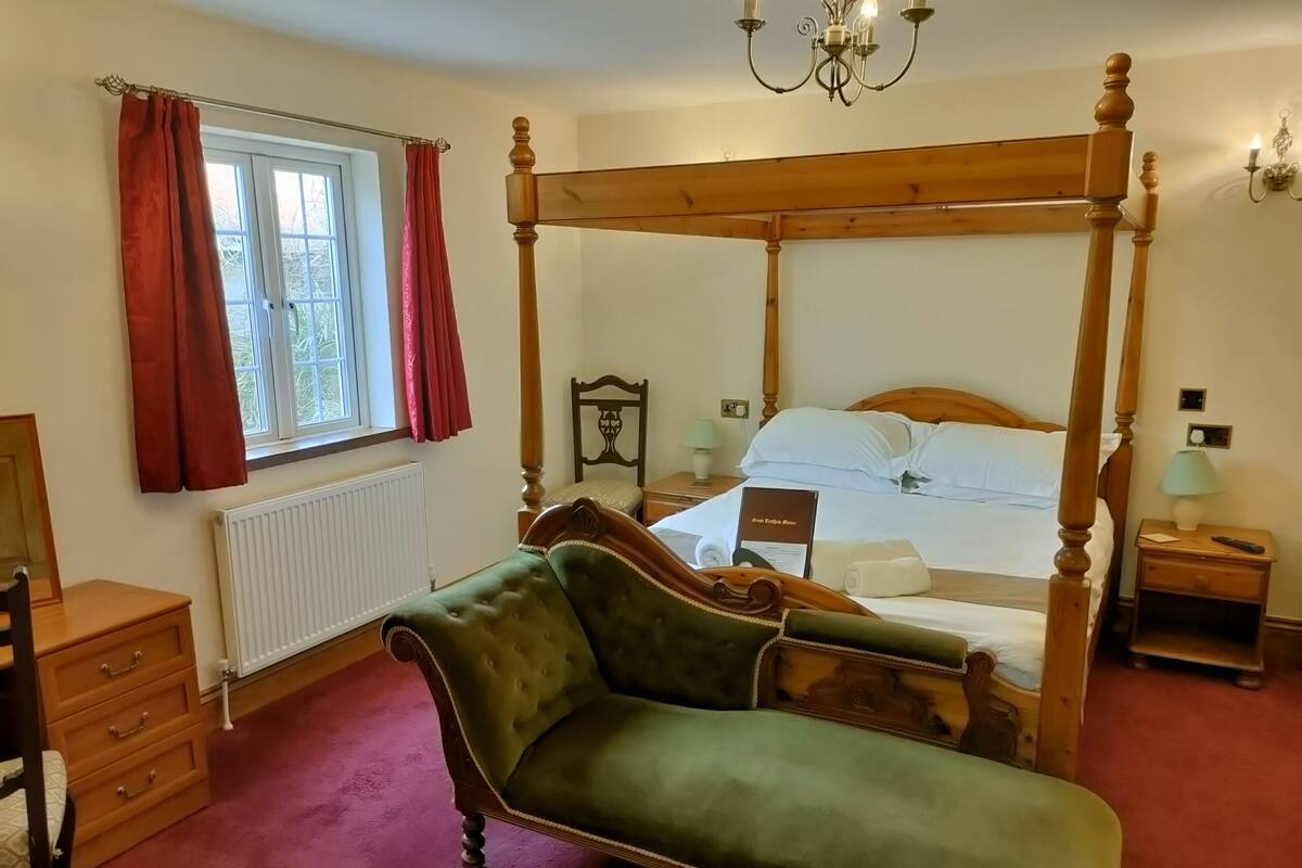 A four-poster bed with a green upholstered headboard, a wooden nightstand with a lamp, a wooden dresser, and a window with red curtains.