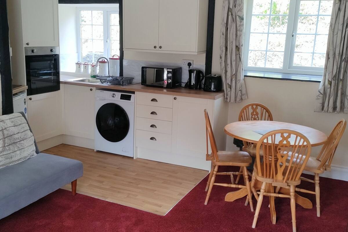 A compact living space with a sofa, a dining table with chairs, a washing machine, a microwave, and a small kitchen area with cabinets and a window.