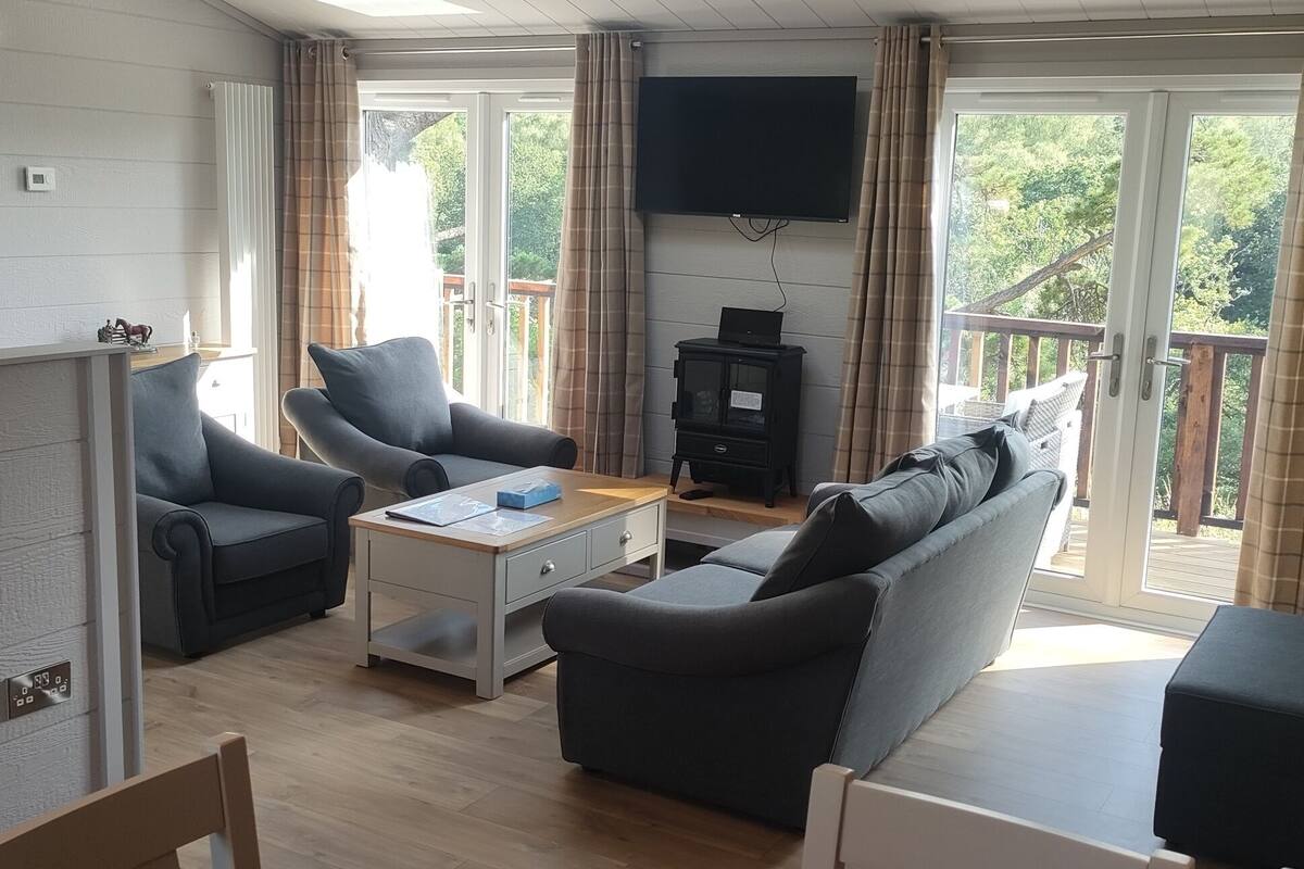 A modern living room with a sofa, armchairs, a coffee table, and a TV. There is a balcony with a view of greenery.