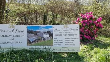 A sign for Peacock Park Holiday Lodges and Caravan Park, Great Trethewn Manor, listing amenities like a bar, restaurant, garden bar, log fires, woodland walks, fishing lake, fish ponds, riding stable, and BBQ area.