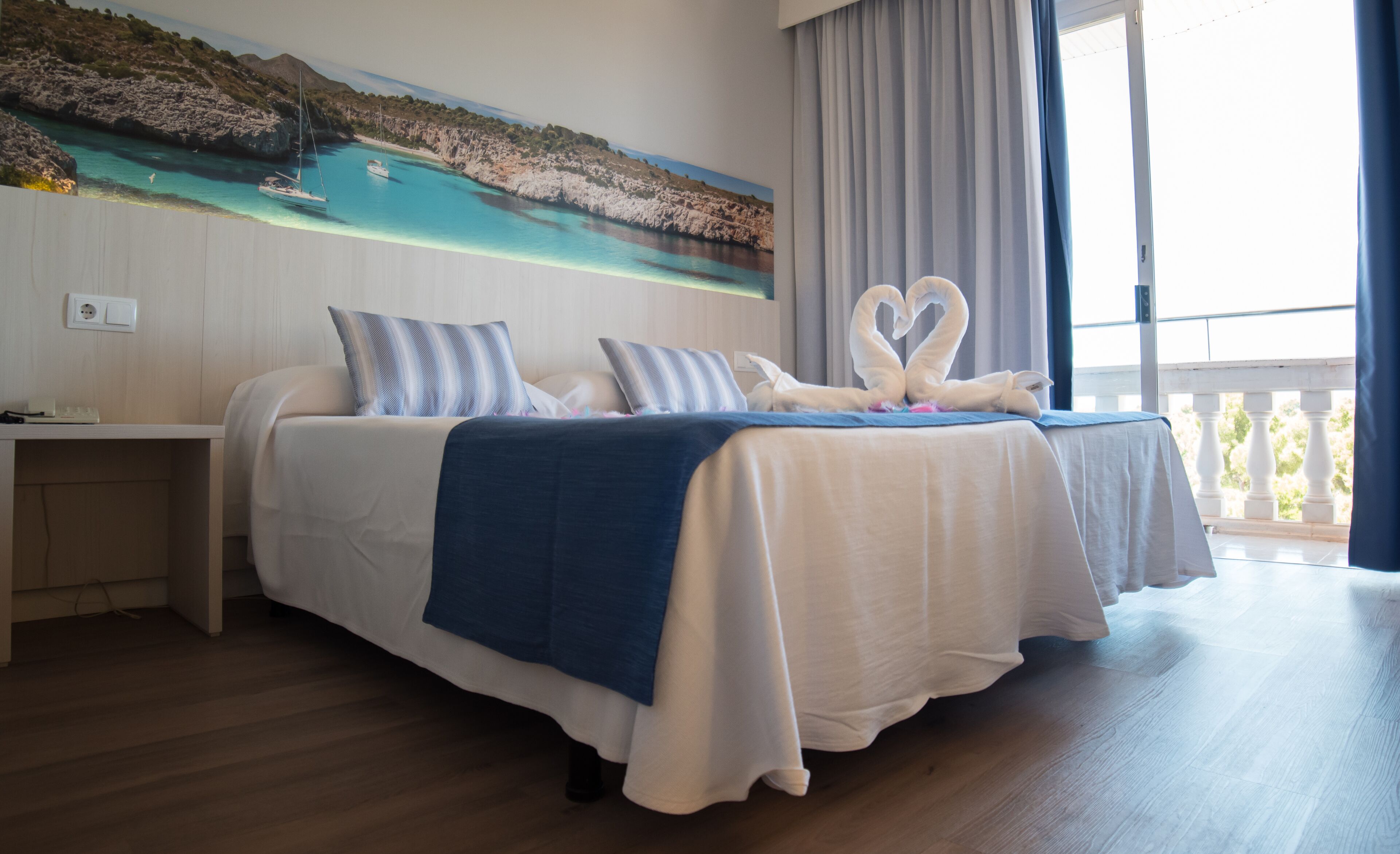 A hotel room with a bed, a nightstand, a painting of a coastal scene, and a balcony with curtains.