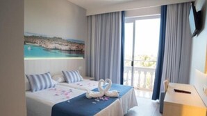 Une chambre d’hôtel avec deux lits, un tableau représentant un paysage côtier, un balcon avec des rideaux et un bureau avec une chaise.