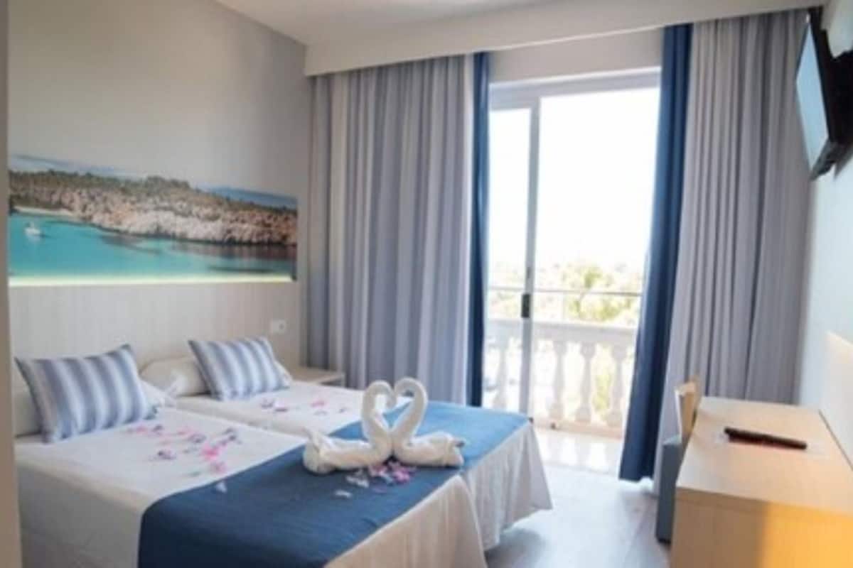 Ein Hotelzimmer mit zwei Betten, einem Gemälde einer Küstenlandschaft, einem Balkon mit Vorhängen und einem Schreibtisch mit Stuhl.