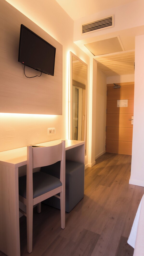 A modern hotel room with a wooden desk and chair, a flat-screen TV mounted on the wall, and a mirror above the desk.