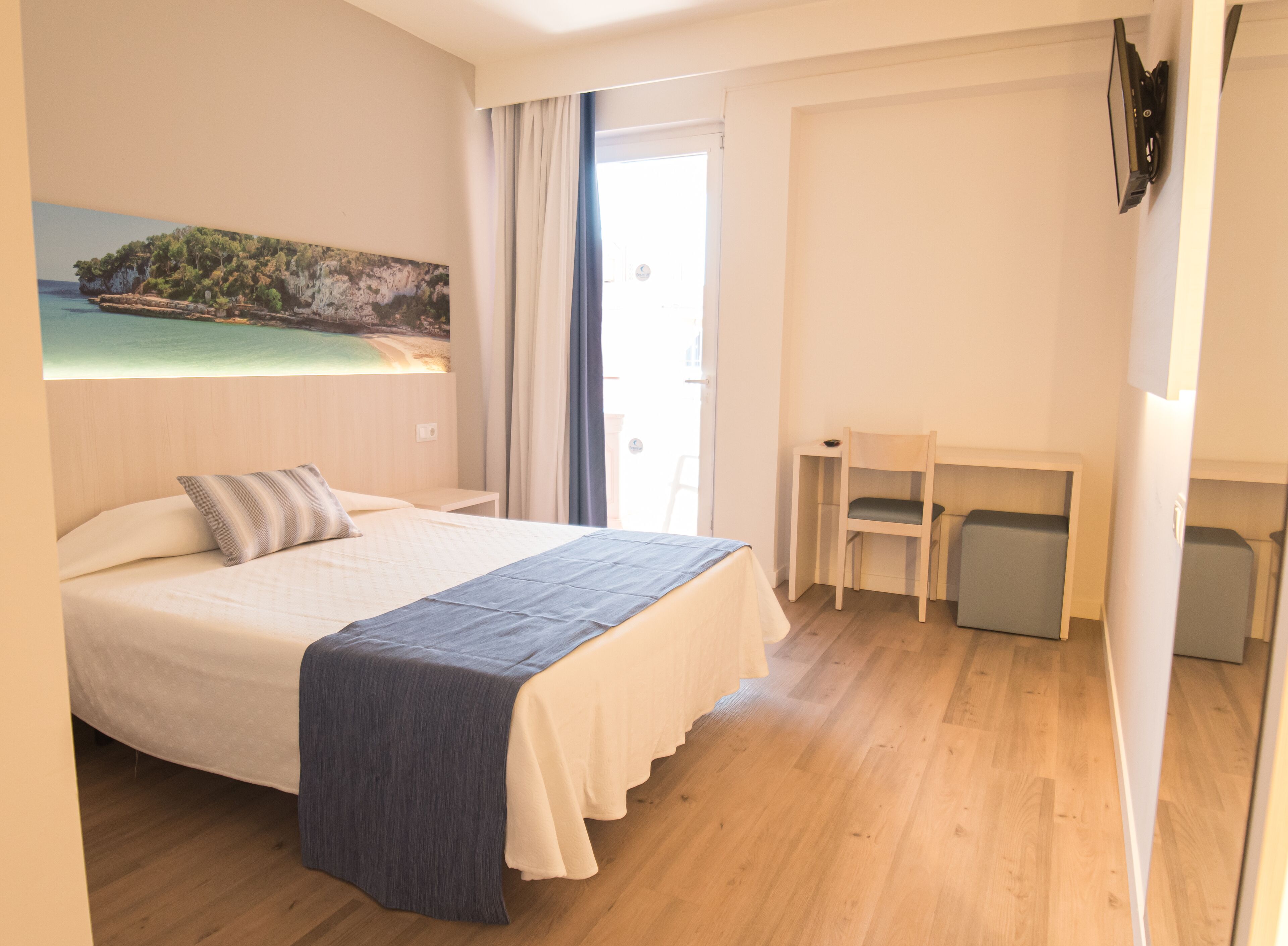 Una camera d'albergo con un letto, una scrivania e una sedia. Appeso al muro, un quadro raffigura una spiaggia.