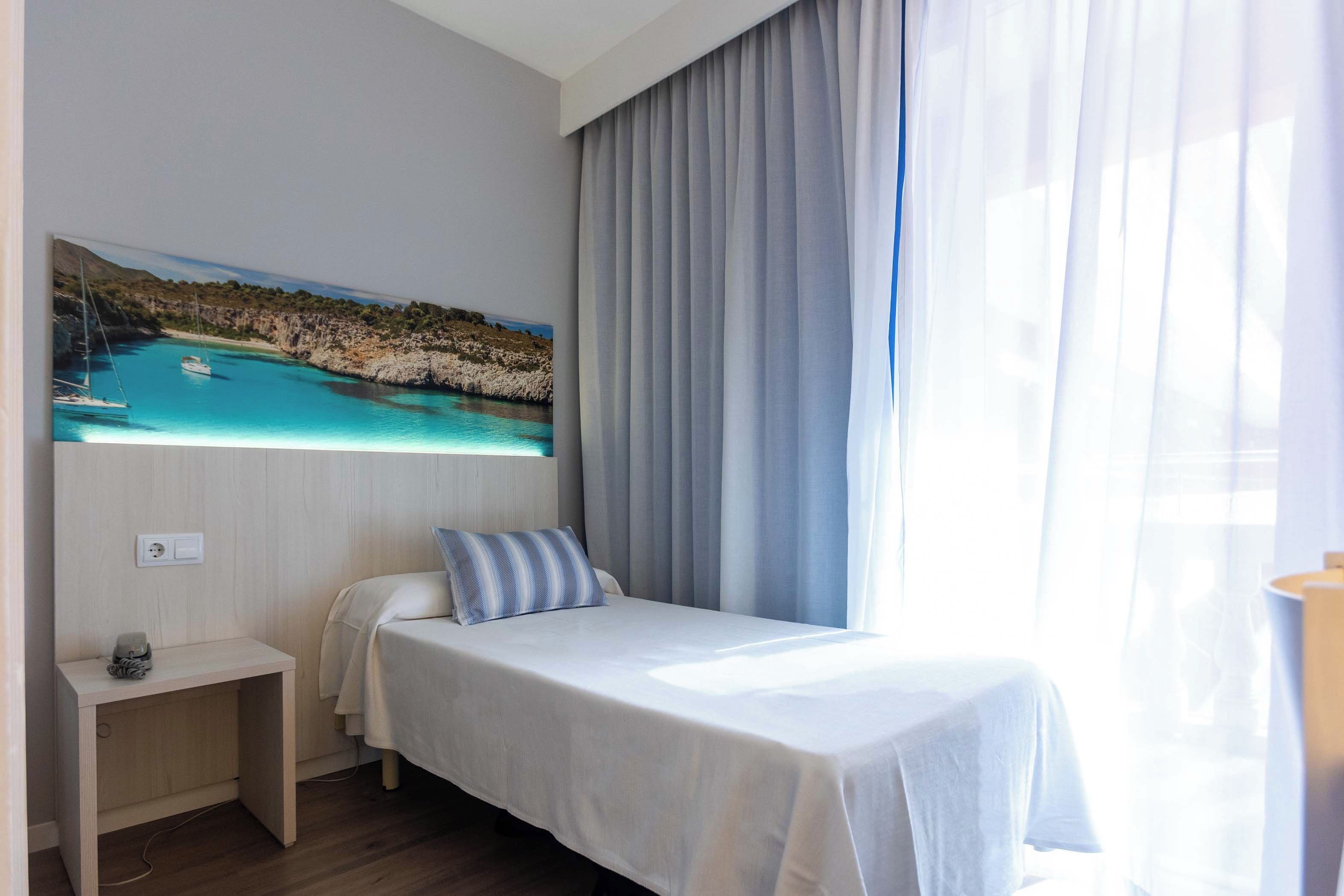 Een hotelkamer met een bed, nachtkastje, schilderij van een kustlandschap en een raam met gordijnen.