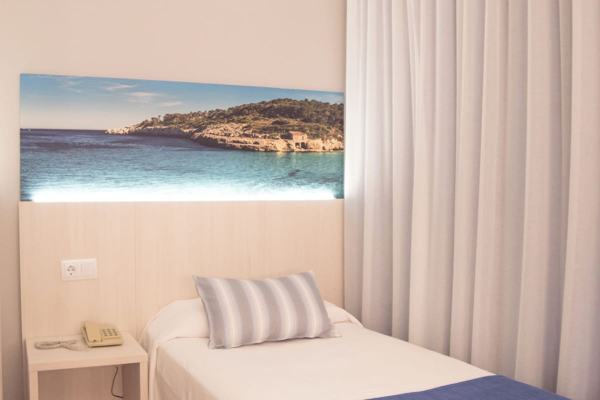 Ein Hotelzimmer mit einem Bett, einem Nachttisch mit Telefon, einem Gemälde einer Küstenlandschaft und einem Fenster mit Vorhängen.