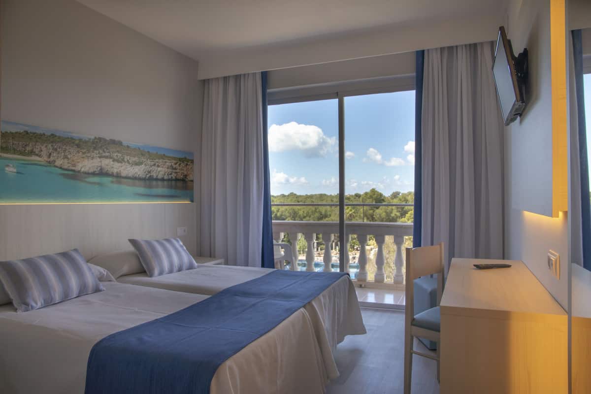 Ein Hotelzimmer mit zwei Betten, einem Schreibtisch und einem Balkon mit Blick auf Meer und Berge.