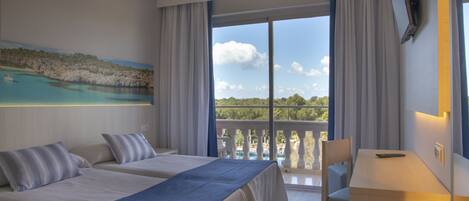 Habitación de hotel con dos camas, un escritorio y un balcón con vistas al mar y a las montañas.