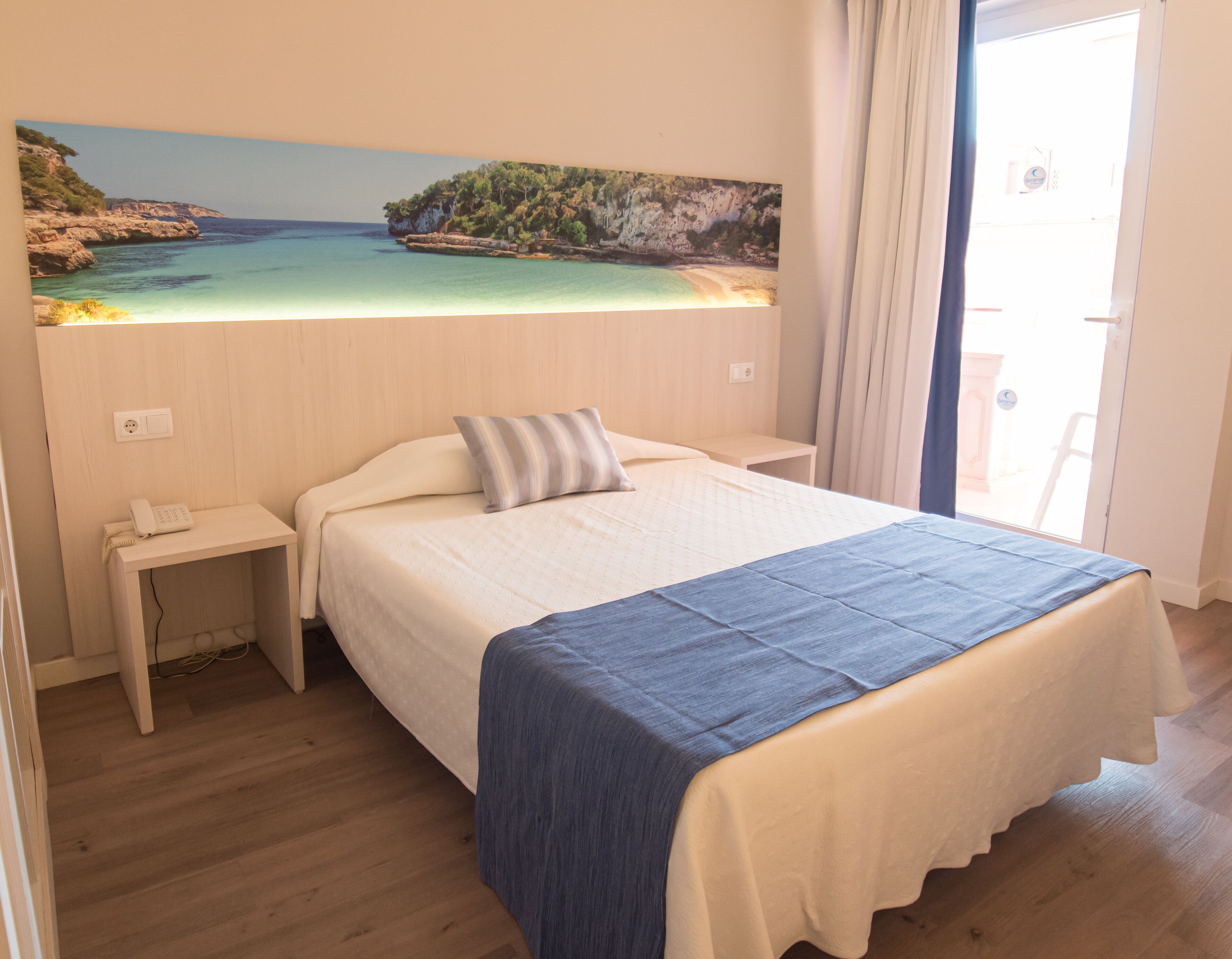 Un dormitorio con cama, mesita de noche, una pintura de paisaje costero y vistas al mar a través de una ventana.
