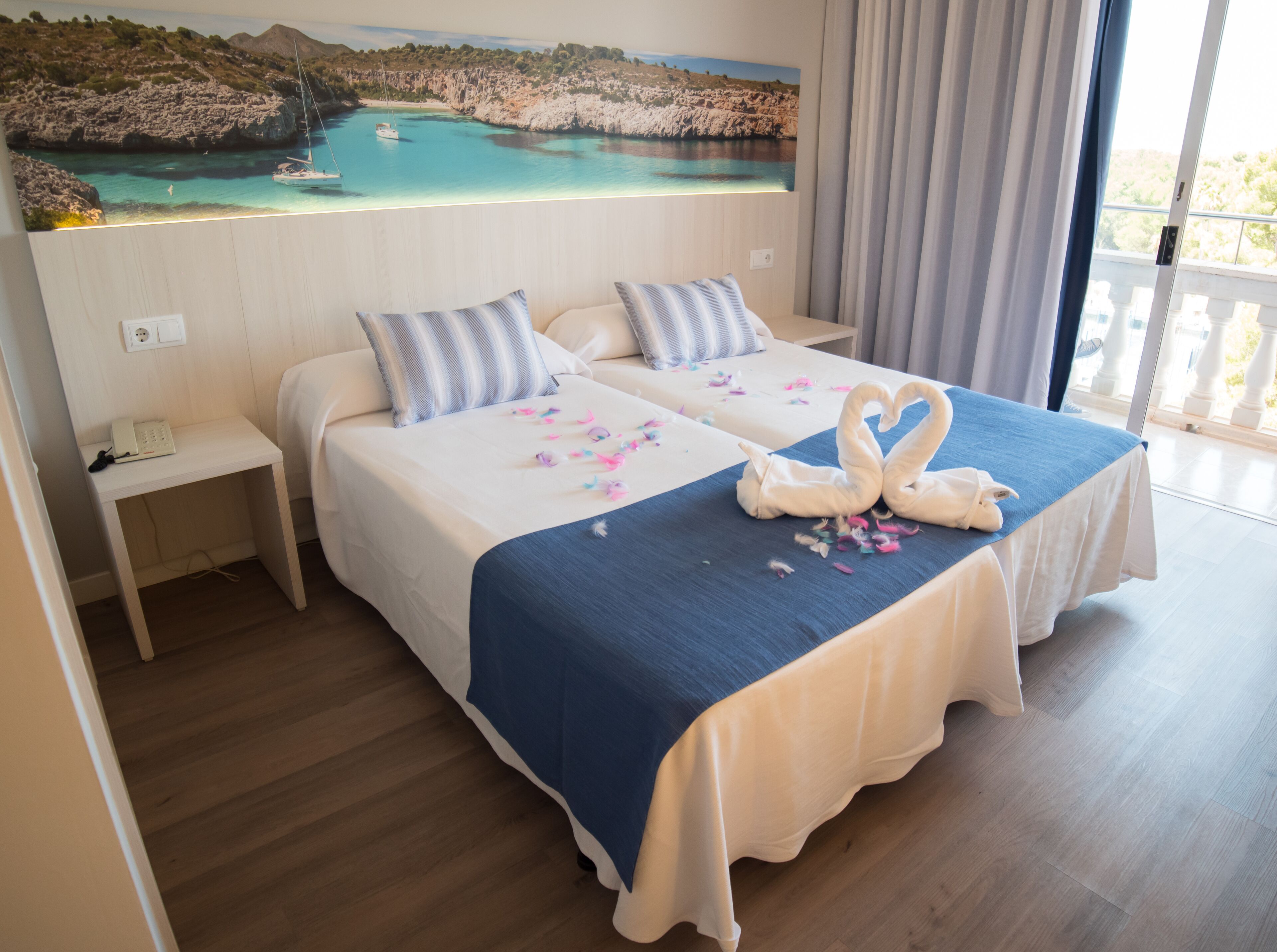 A hotel room with two beds, a painting of a coastal scene, and a view of the sea through the window.
