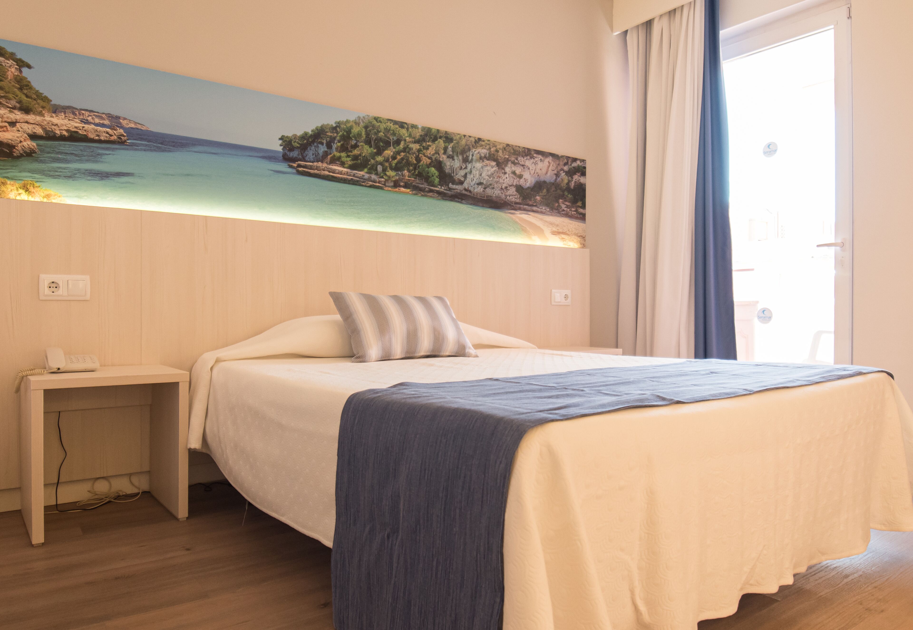 A hotel room with a bed, a nightstand, a painting of a beach scene, and a door to the right.