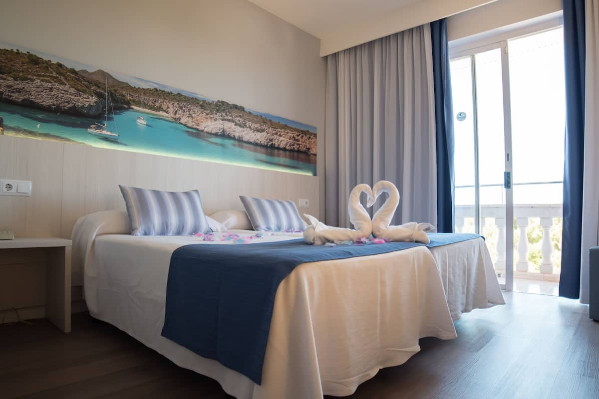 Ein Hotelzimmer mit einem großen Bett, einem Gemälde einer Küstenlandschaft und einem Balkon mit Vorhängen.