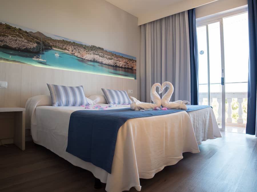Habitación de hotel con una cama grande, un cuadro de paisaje costero y un balcón con cortinas.