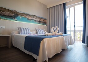 Habitación de hotel con una cama grande, un cuadro de paisaje costero y un balcón con cortinas.