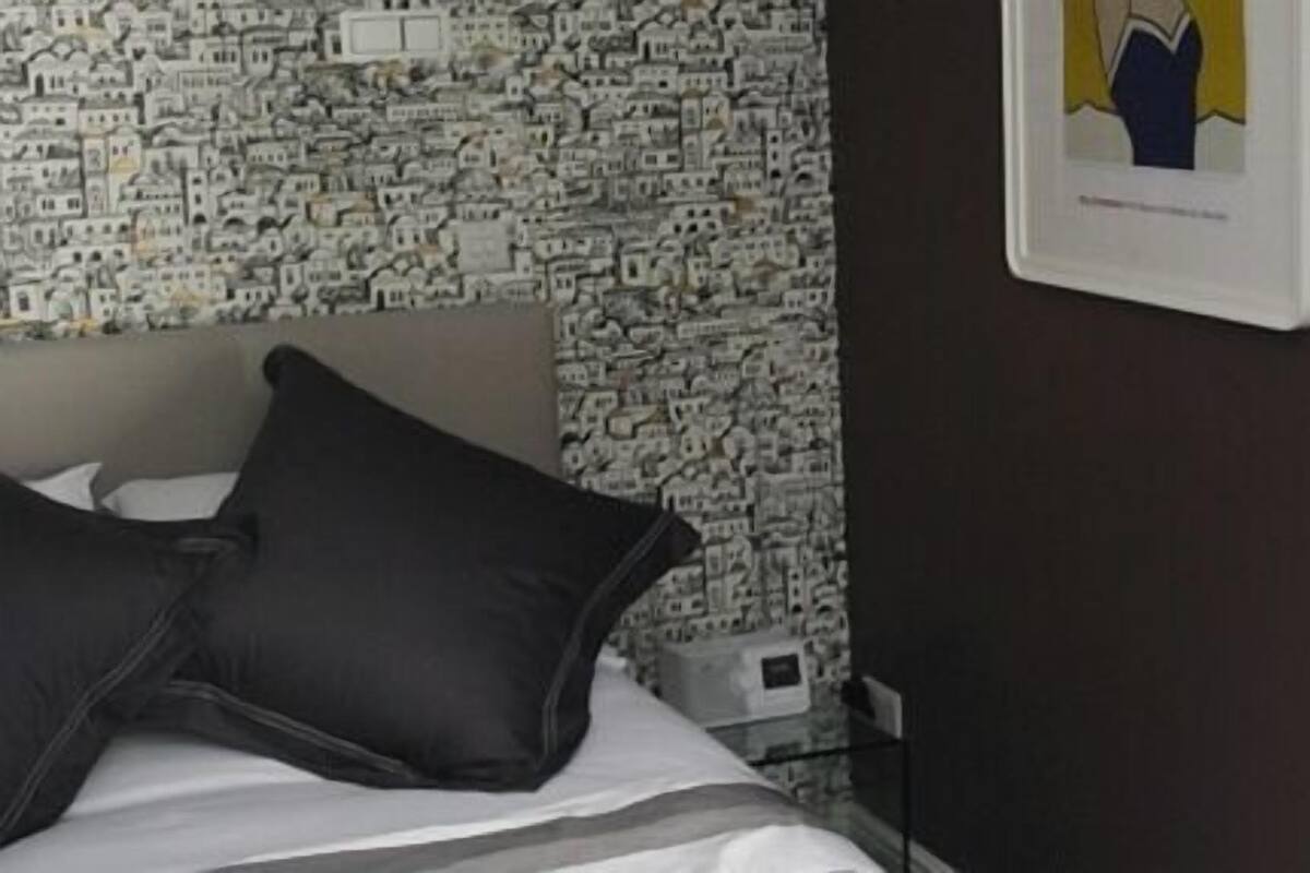 A hotel room with a patterned wallpaper, a bed with dark pillows, and a framed picture on the wall.