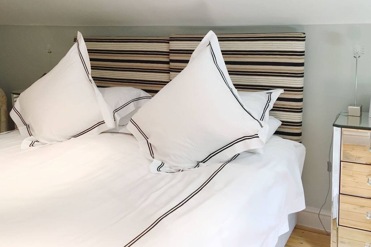 A neatly made bed with white linens and striped headboard, a wooden dresser with drawers, and a floral-patterned chair.