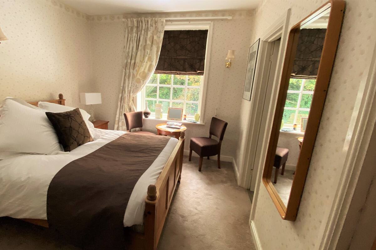 A neatly made bed with white and brown linens, a bedside table with a lamp, a chair, and a window with curtains.