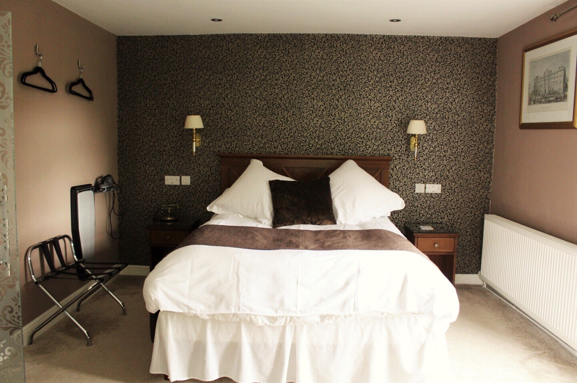 A neatly made bed with white bedding and a brown accent, a bedside table with a lamp, a chair, and a wall-mounted coat rack.