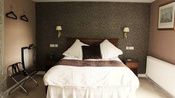 A neatly made bed with white bedding and a brown accent, a bedside table with a lamp, a chair, and a wall-mounted coat rack.