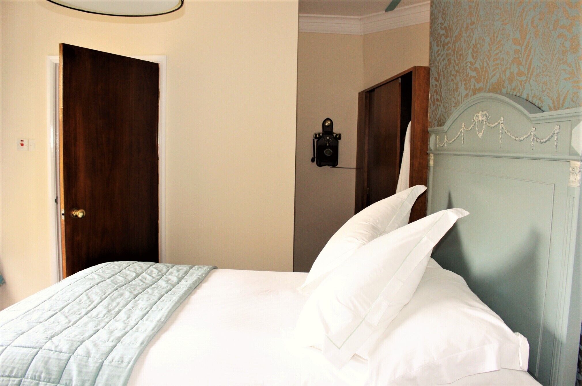 A neatly made bed with a quilted blanket and white pillows, a wooden door, and a wall with a floral wallpaper pattern.