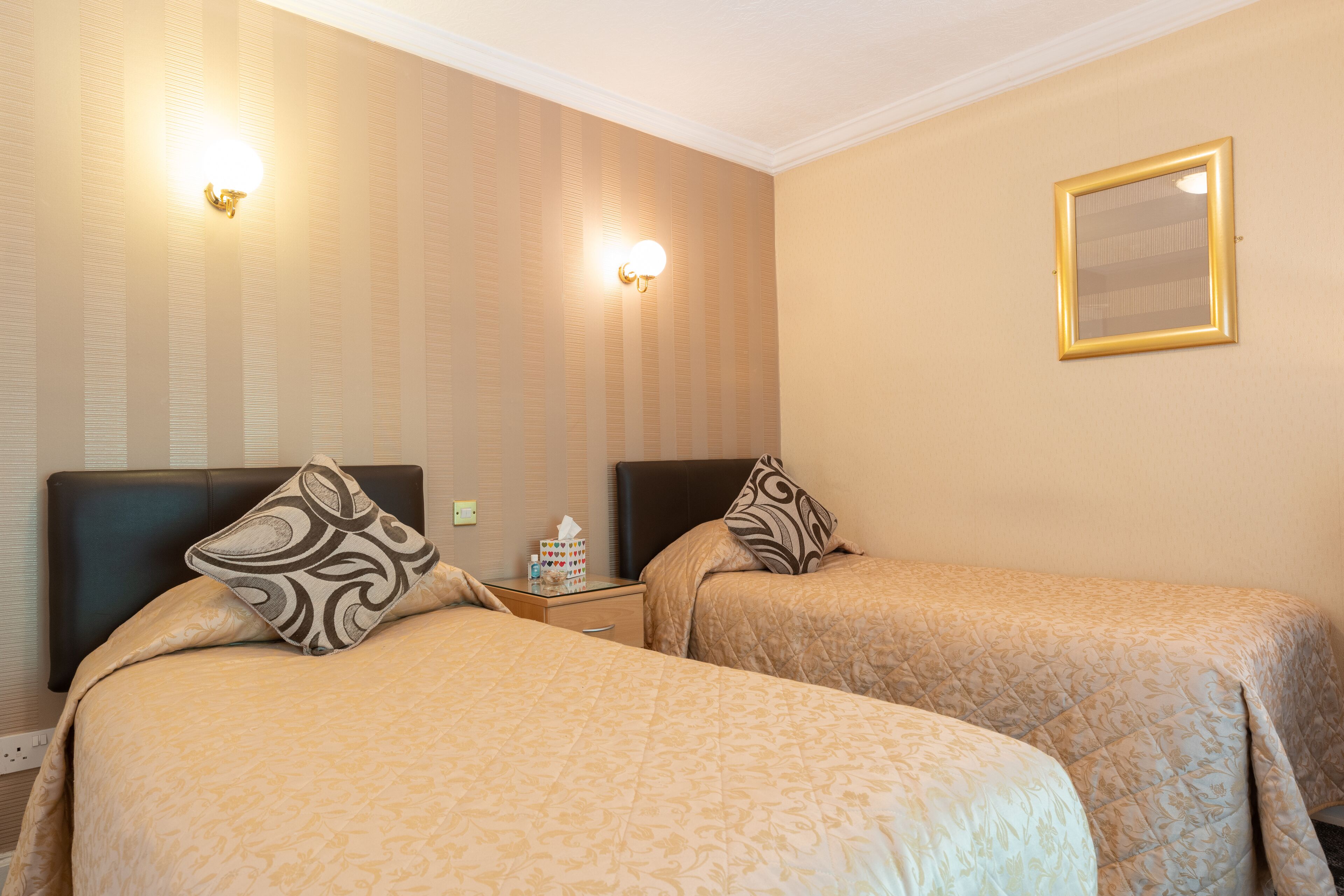 A hotel room with two beds, each with a headboard, a bedside table with a lamp, and a wall-mounted mirror.